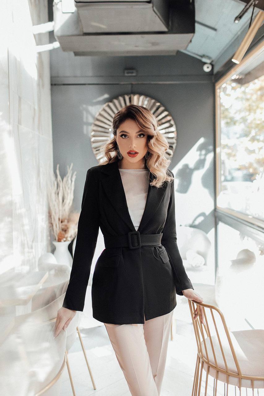 Woman in a black blazer and white top standing in a modern interior setting.
