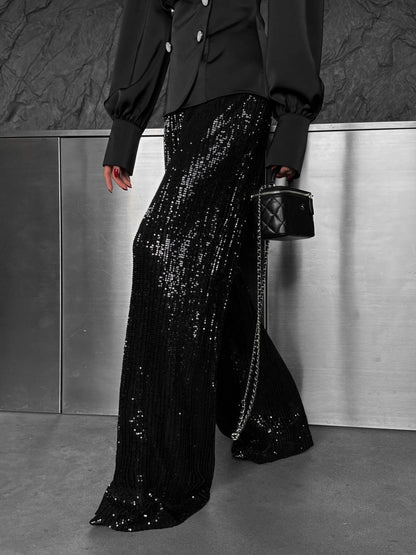 Person wearing black sequin pants and a black jacket with a small black handbag against a metallic wall.