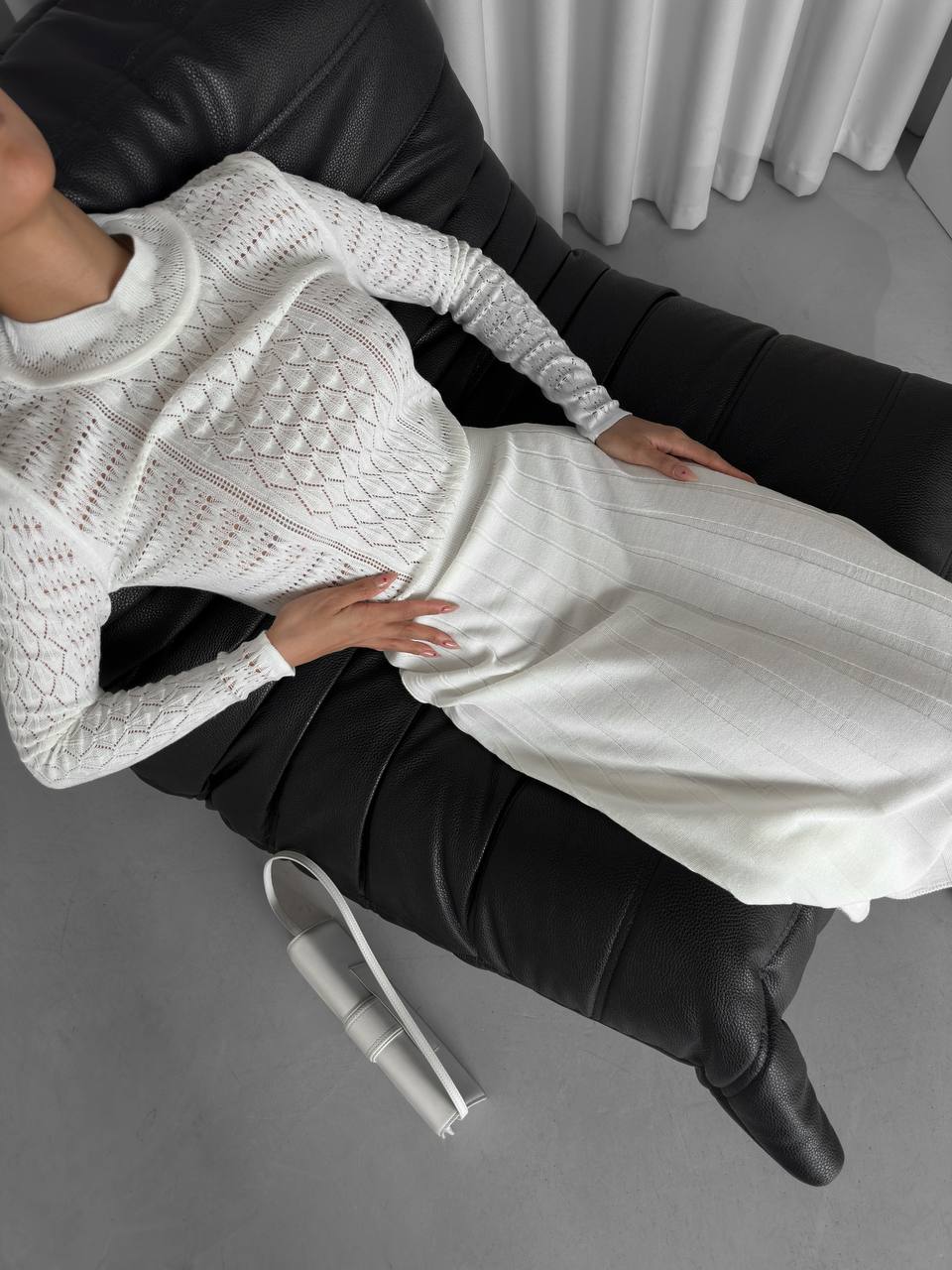 Person wearing a white textured sweater and black pants sitting on a gray floor.