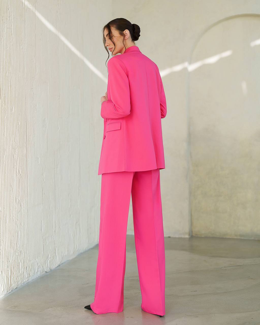 Person wearing a bright pink suit standing in a minimalistic room with white walls.