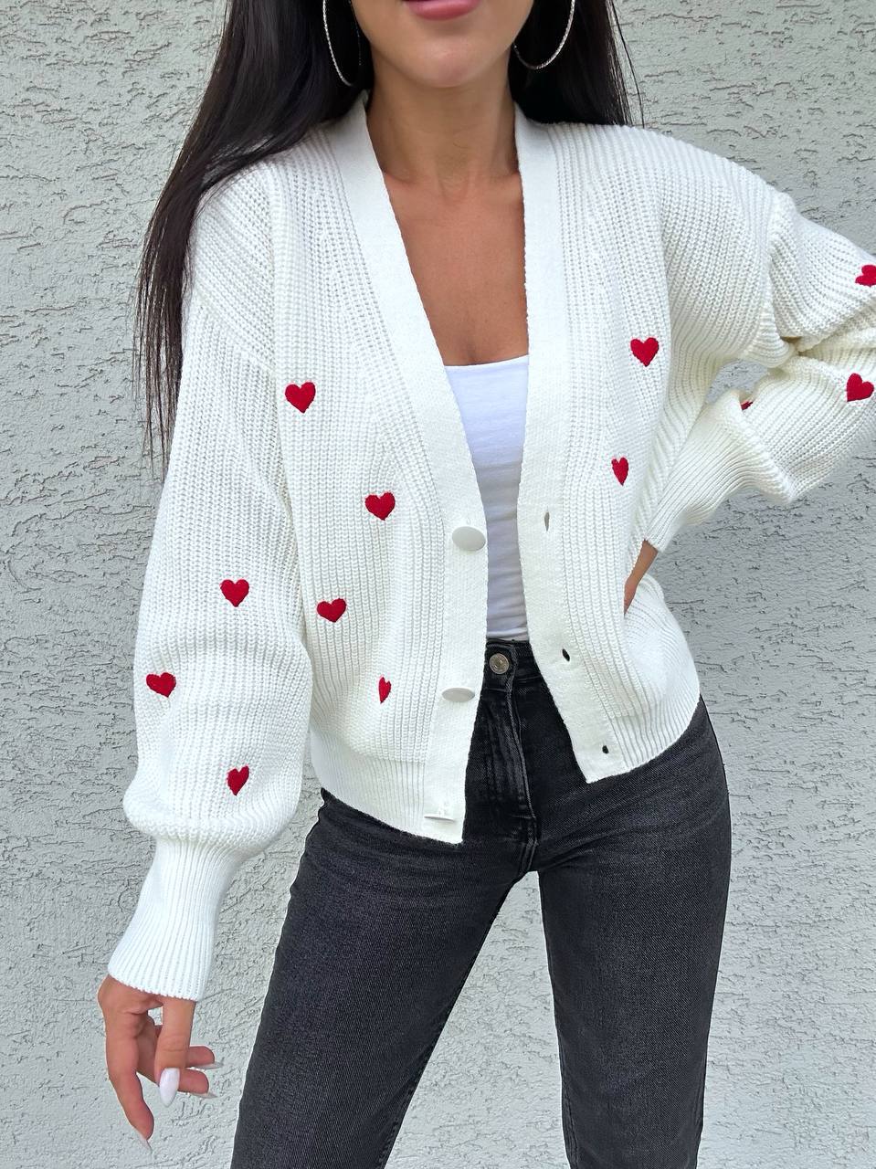 White cardigan with red heart patterns worn by a person against a gray background