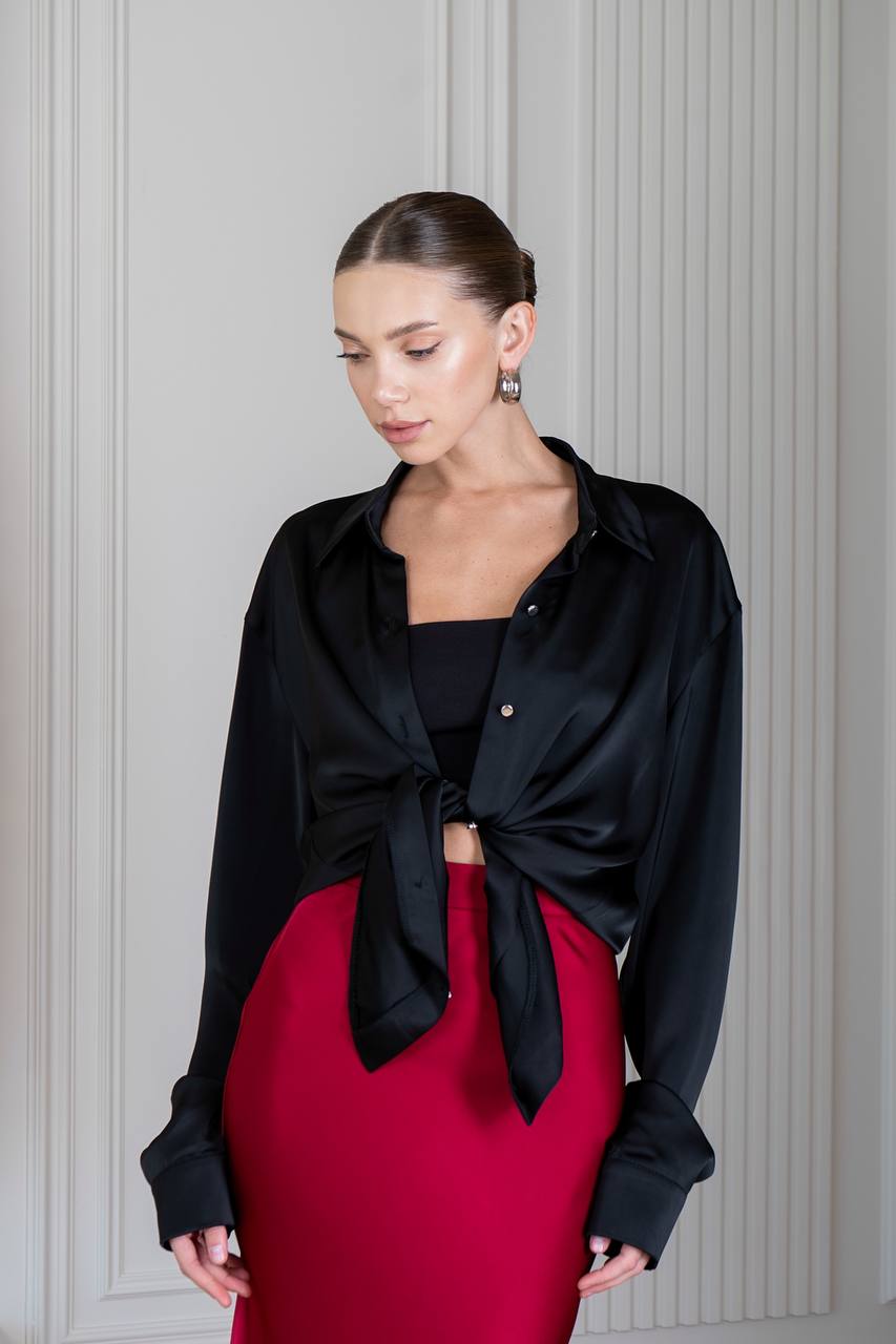 Woman wearing a black tied blouse over a red skirt against a gray paneled wall.