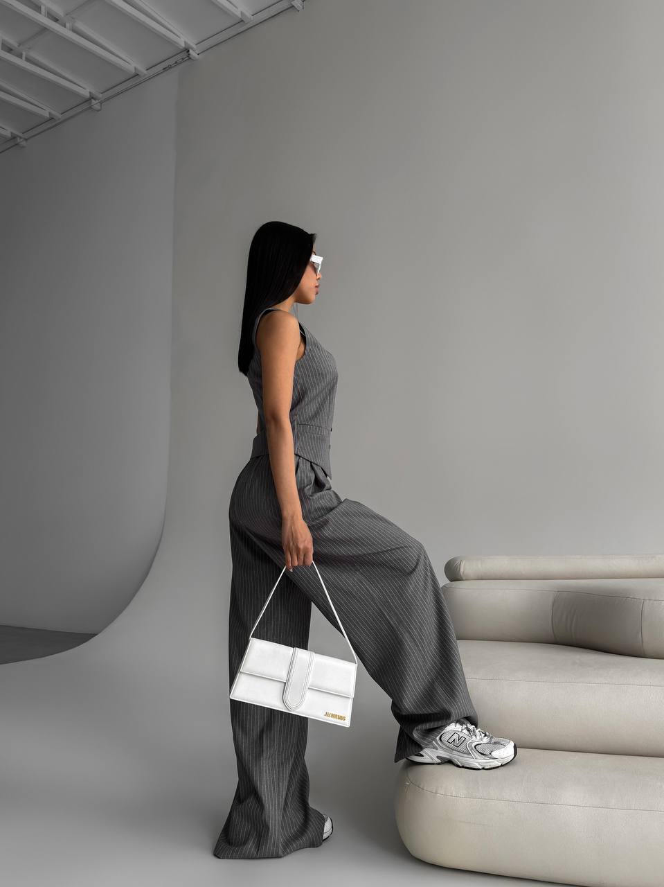 Woman in a gray outfit holding a white handbag against a minimalistic background