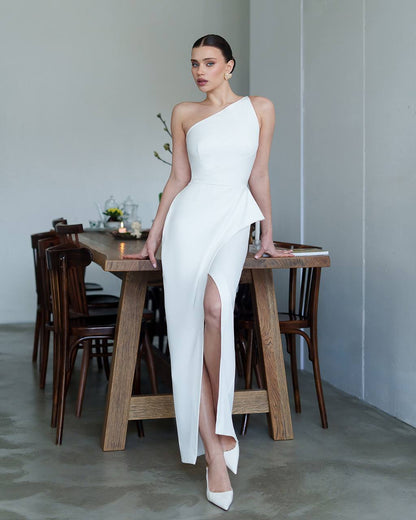 Woman in a white one-shoulder dress standing by a wooden table indoors.