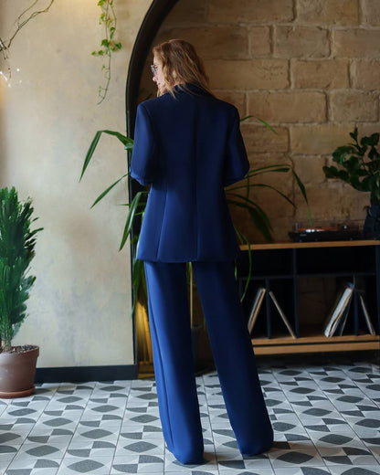 Woman in a blue suit standing in a room with plants and a brick wall.
