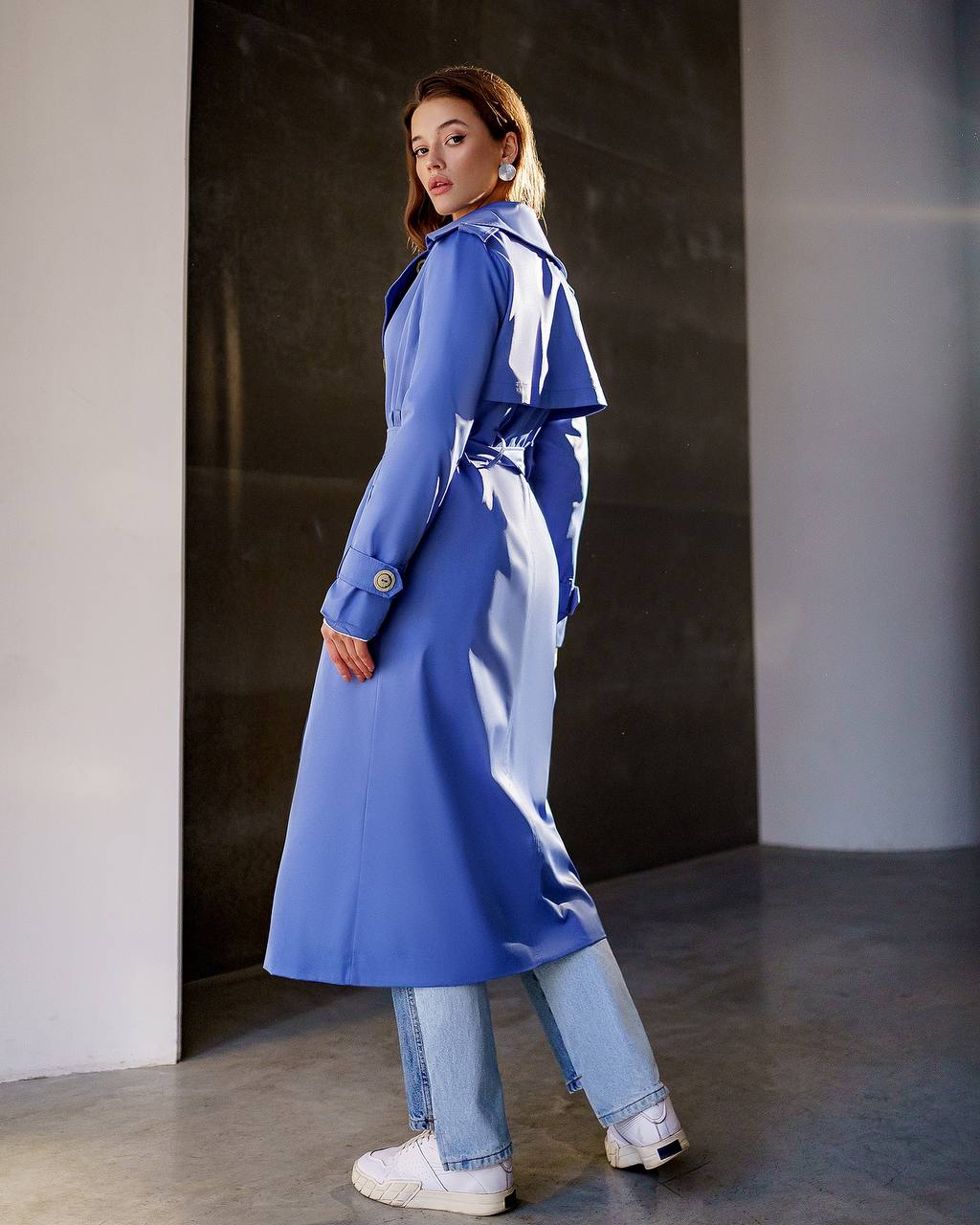Woman wearing a blue trench coat and jeans standing against a dark wall.