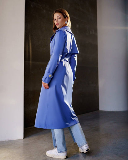Woman wearing a blue trench coat and jeans standing against a dark wall.