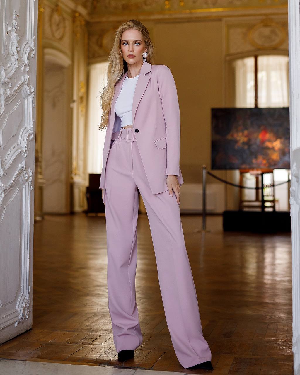 Woman in a pink suit standing in an elegant room with ornate decor.