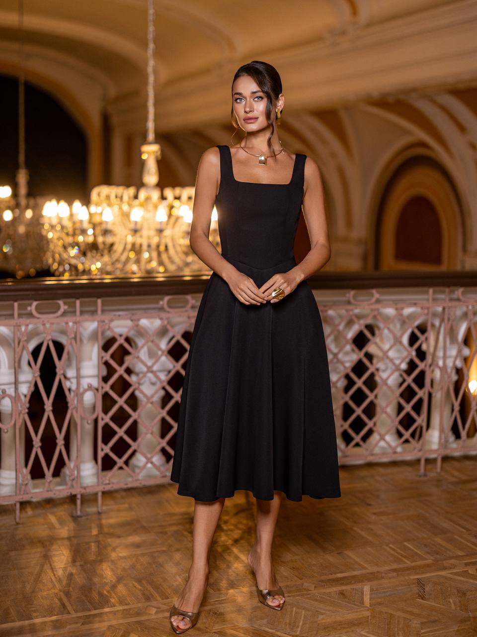 Woman in a black dress standing in an elegant indoor setting with chandeliers.