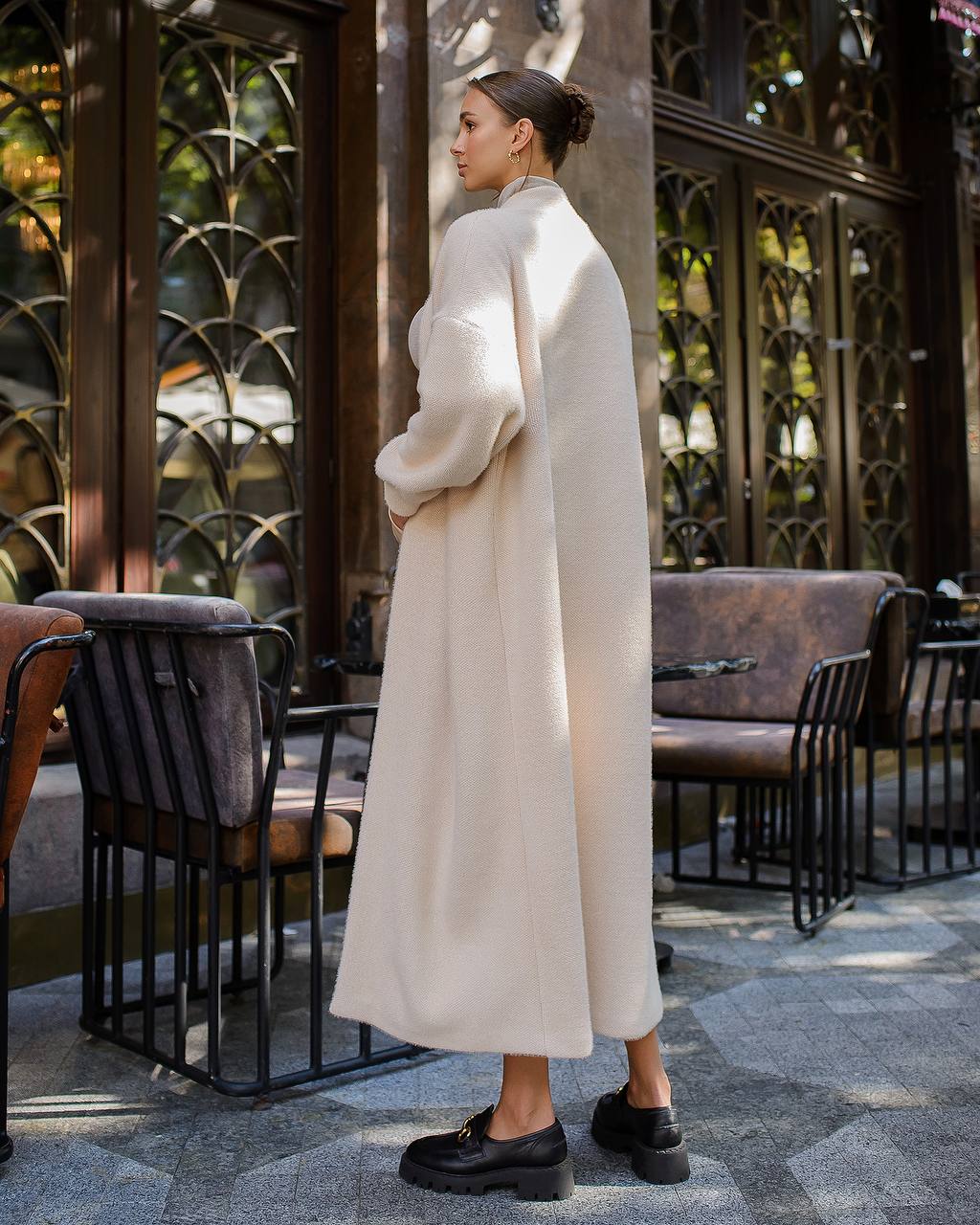 Woman in a long white coat standing in an outdoor setting with decorative metalwork.