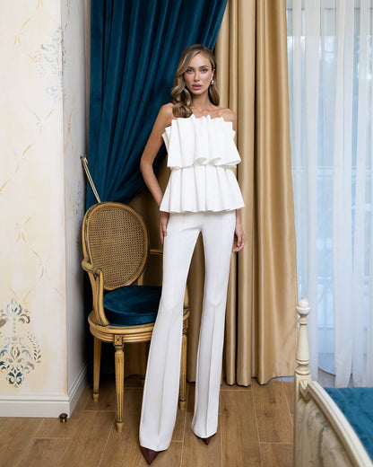 A woman models a white off-the-shoulder ruffled top paired with matching white wide-leg trousers in an elegant room with blue and gold curtains.