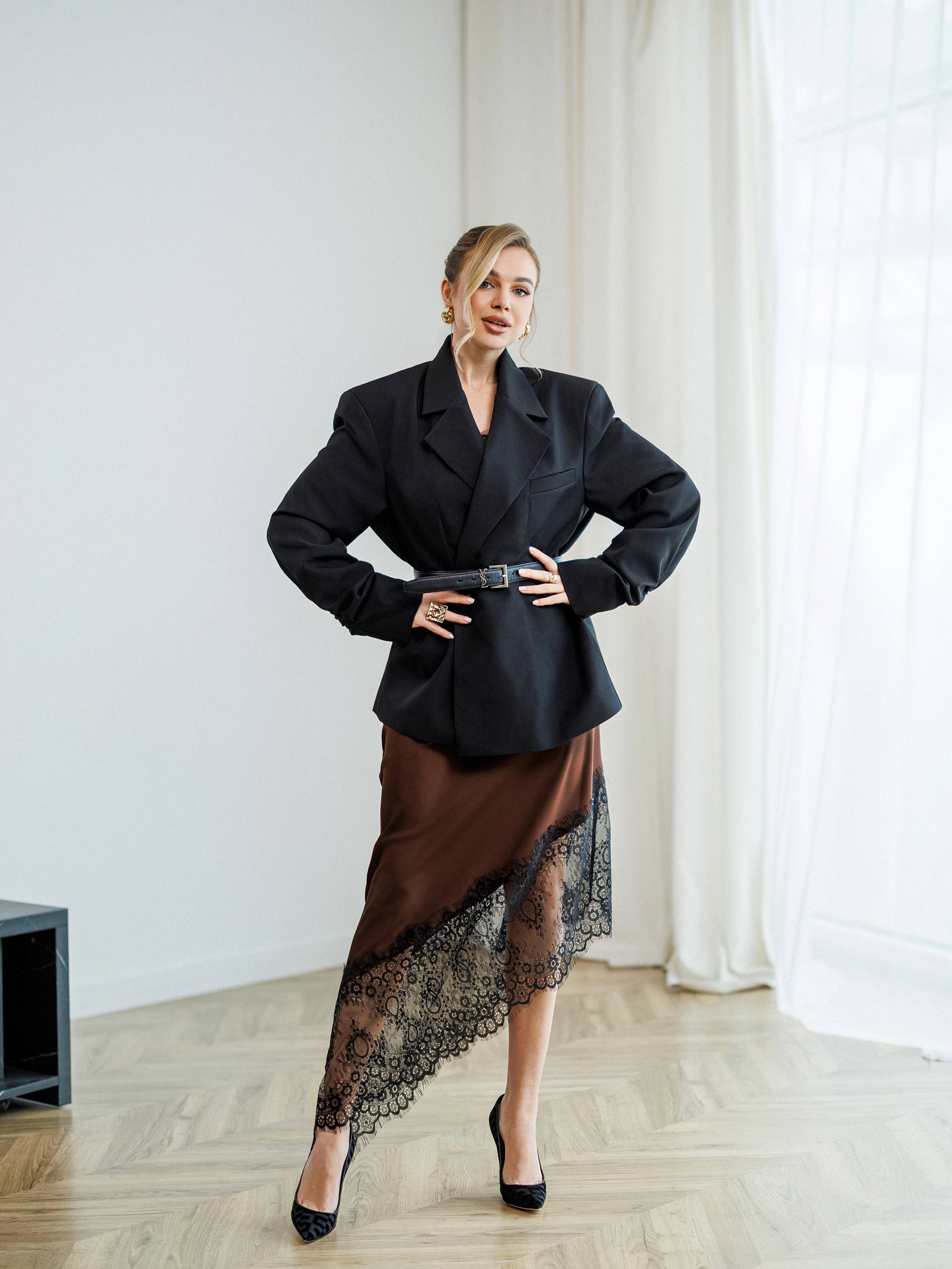 Woman in a black blazer and brown dress standing in a room with white walls and curtains.