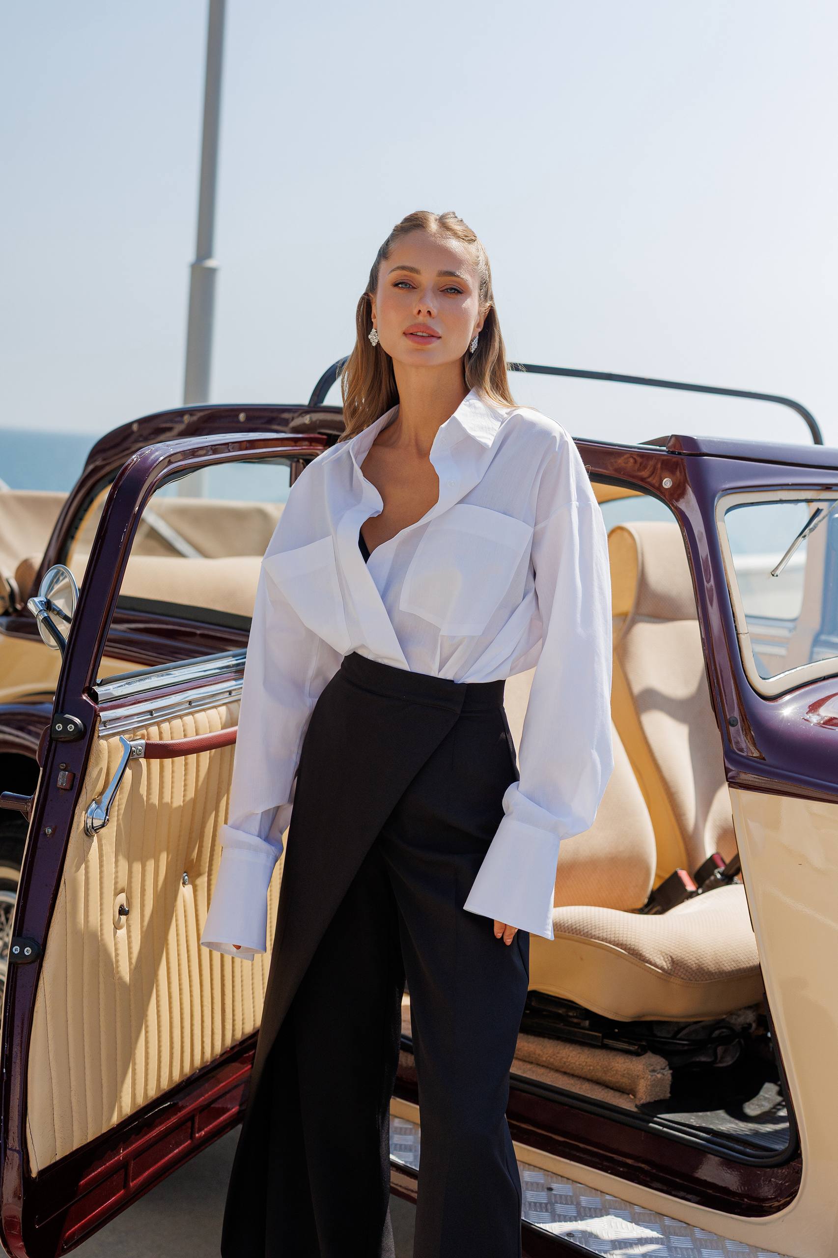 Woman in a white blouse and black pants standing next to an open car door.