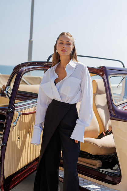 Woman in a white blouse and black pants standing next to an open car door.