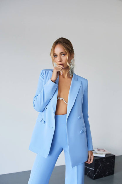 Woman wearing a light blue suit with a white top underneath, standing against a plain background.