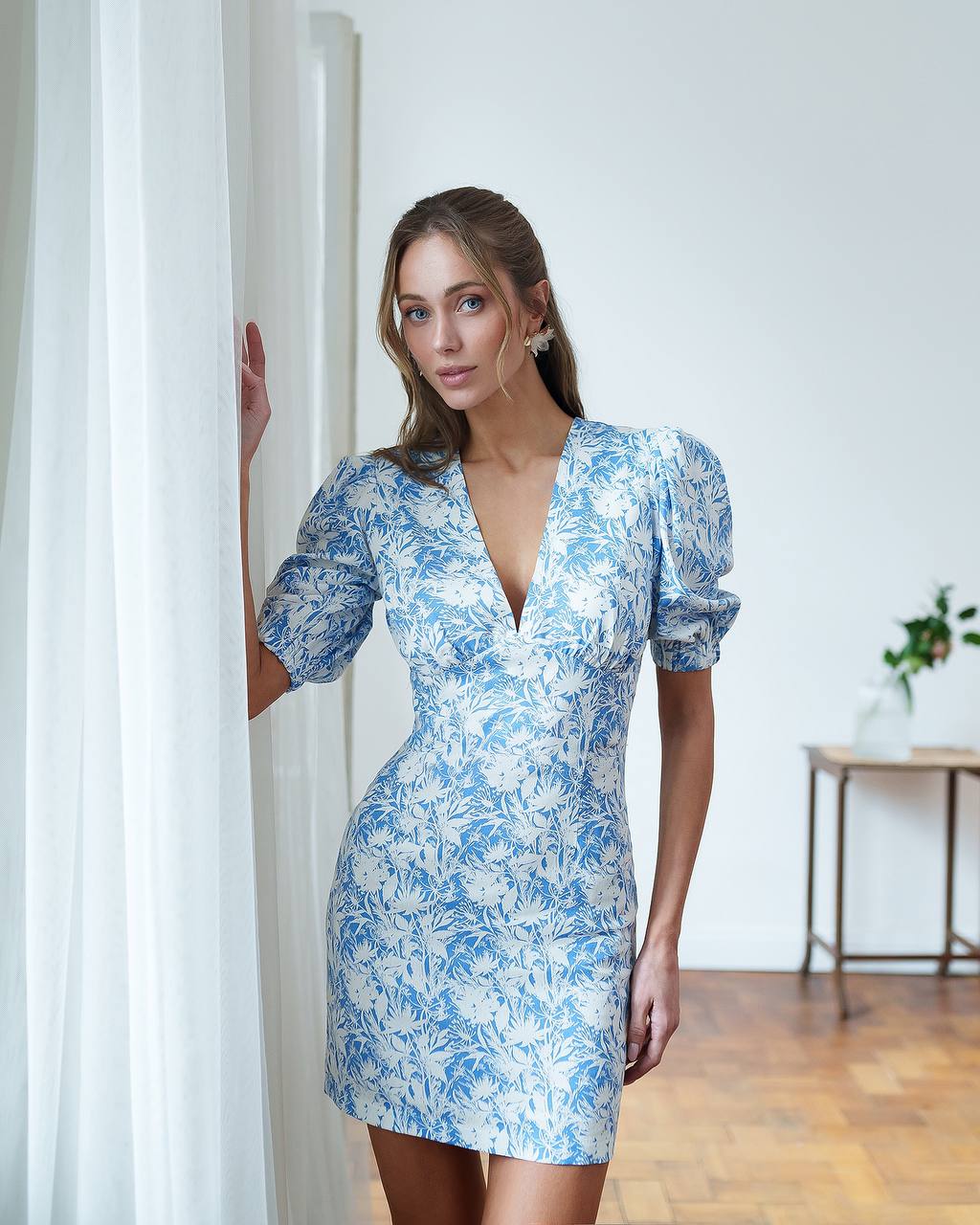 Woman wearing a blue floral dress standing in a room with white walls and wooden flooring.