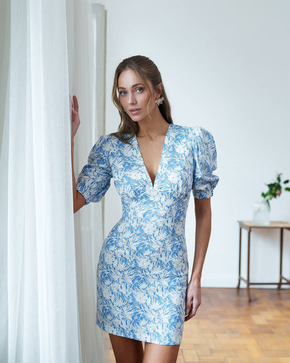 Woman wearing a blue floral dress standing in a room with white walls and wooden flooring.