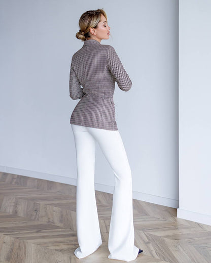 Woman wearing a patterned blazer and white pants standing against a plain wall.