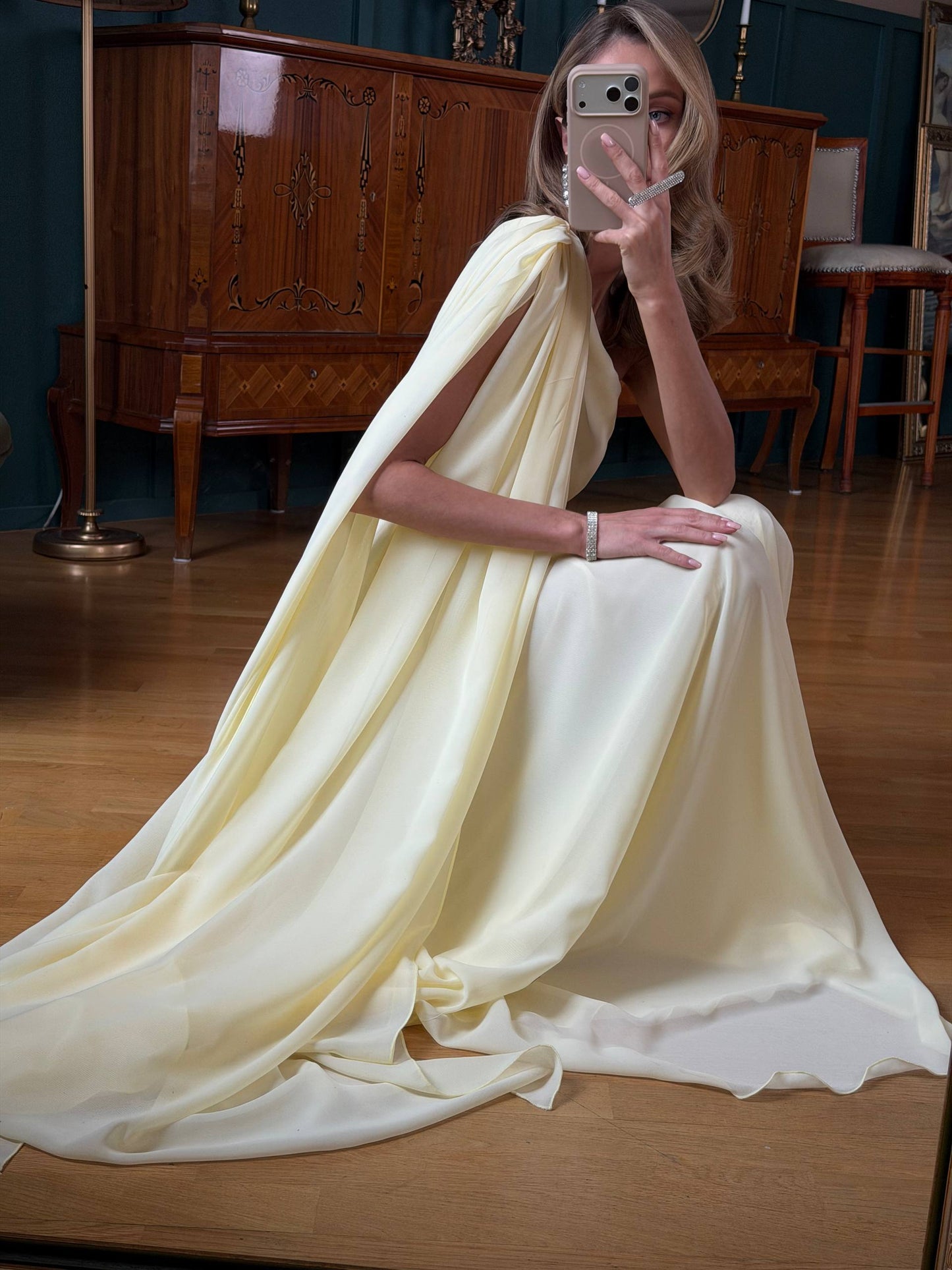 Woman in a flowing white dress sitting on a wooden floor in a room with wooden furniture.