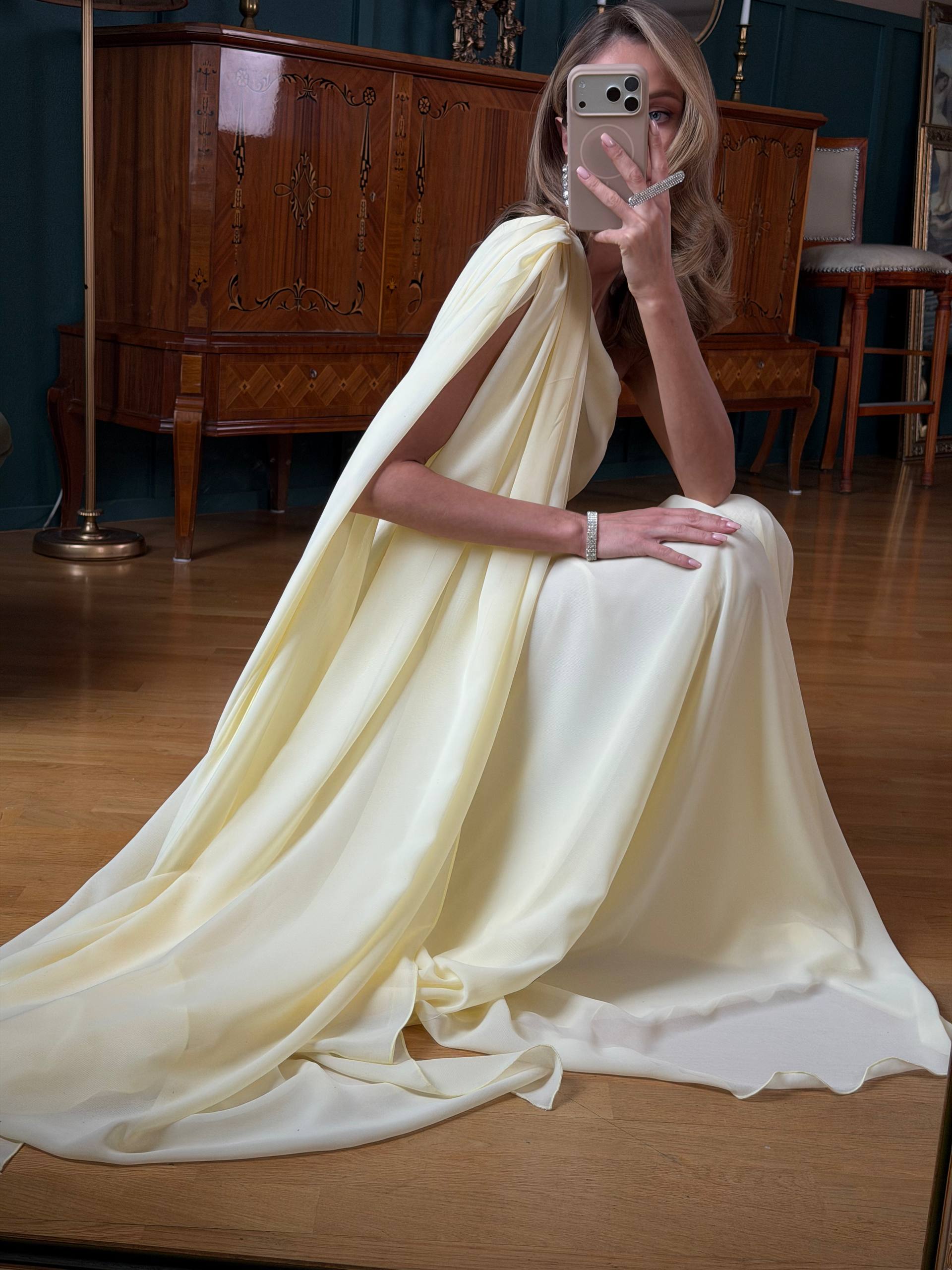 Woman in a flowing white dress sitting on a wooden floor in a room with wooden furniture.