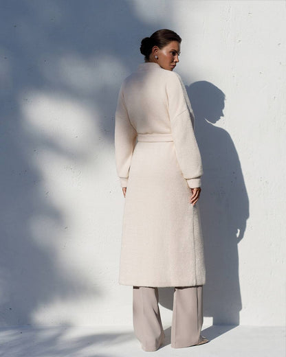 Woman wearing a long beige coat against a white wall