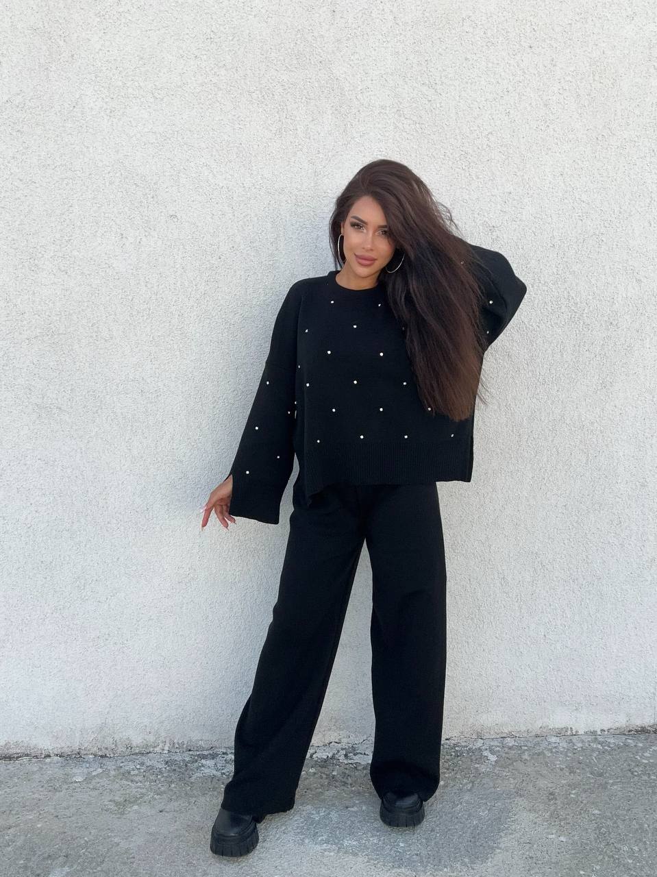Woman wearing a black outfit with white embellishments against a light gray wall.