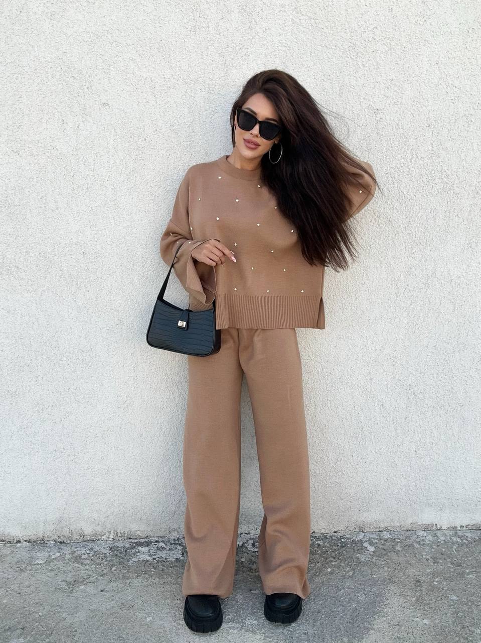 Woman in a brown outfit holding a black handbag against a white wall.