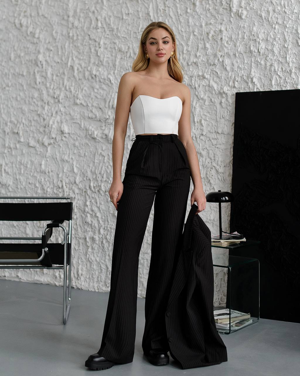 Woman wearing a white strapless top and black high-waisted pants in a minimalistic room.