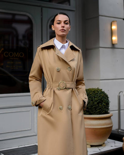 Woman wearing a beige trench coat standing in front of a building entrance.