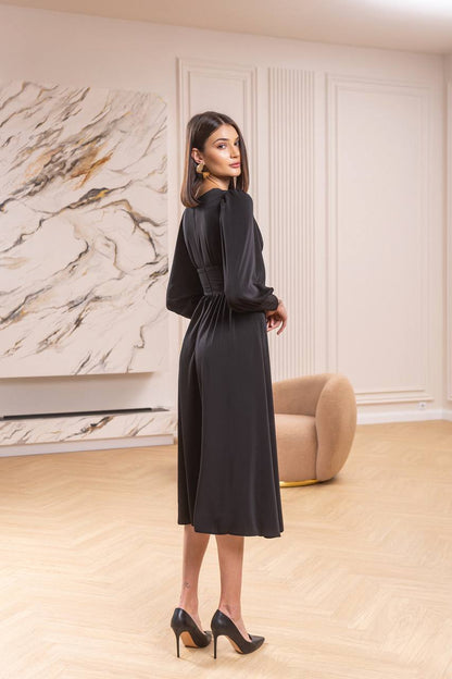 Woman in a black dress standing in a stylish room with marble wall and beige armchair.