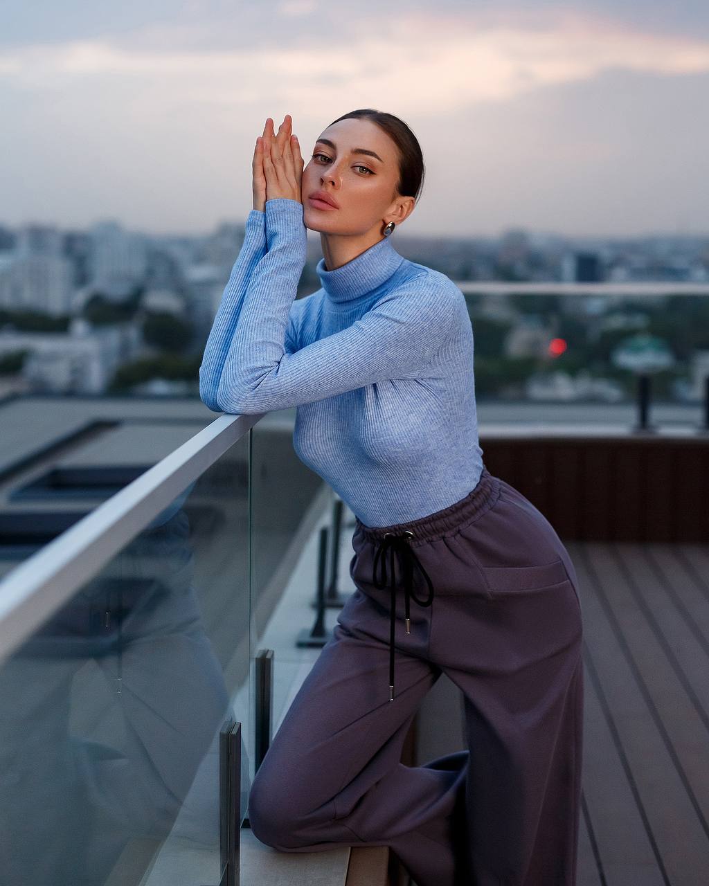Woman in a light blue sweater and purple pants standing on a balcony with a cityscape background.