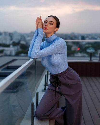 Woman in a light blue sweater and purple pants standing on a balcony with a cityscape background.