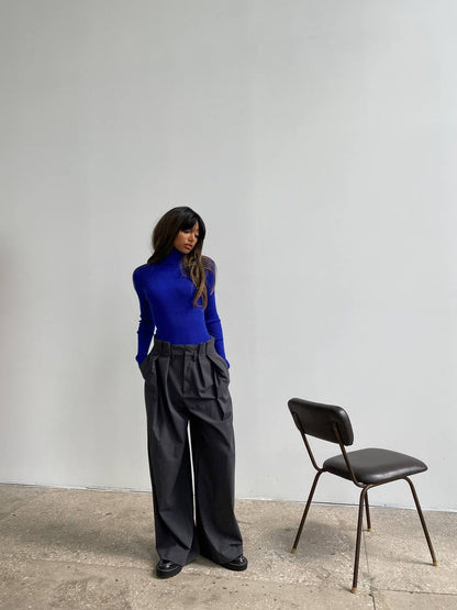 Woman in a blue top and black pants standing next to a chair against a plain wall.