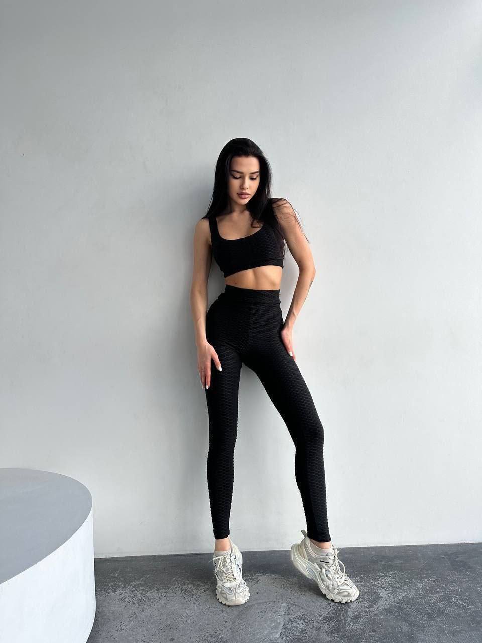 Woman wearing a black athletic outfit standing against a gray wall.