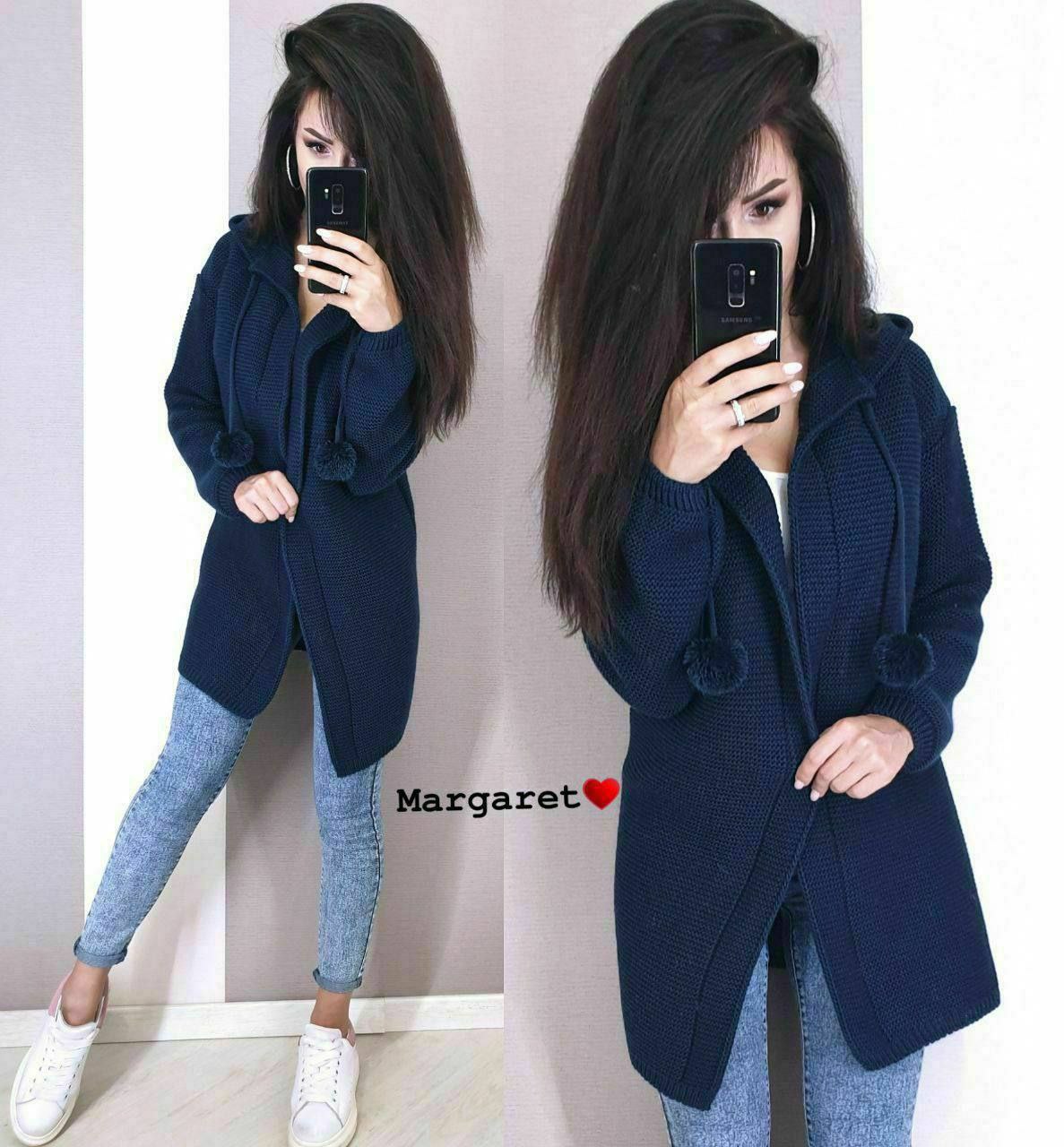 Woman wearing a navy blue coat and blue jeans, holding a phone, with a plain background.