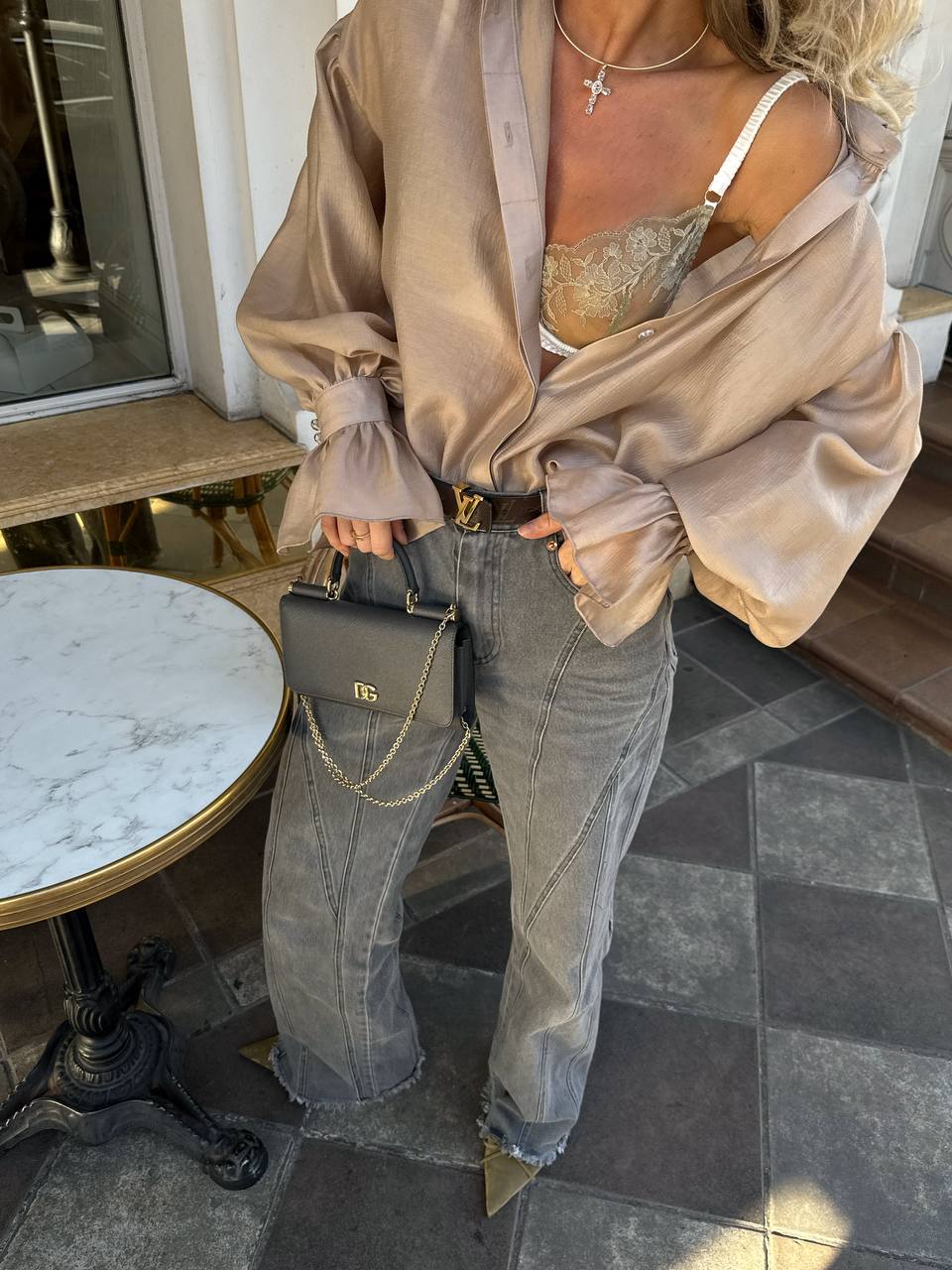 Person wearing a beige satin shirt, bra, and jeans holding a black handbag on a sidewalk.