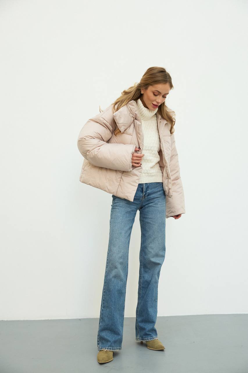 Woman wearing a light pink puffer jacket, white sweater, blue jeans, and brown shoes on a plain background