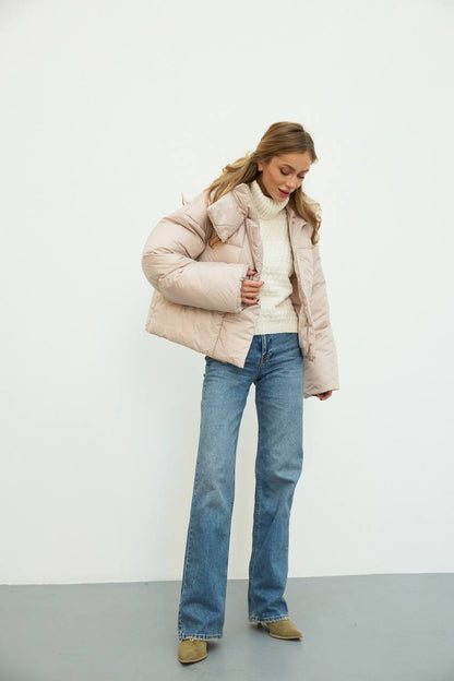 Woman wearing a light pink puffer jacket, white sweater, blue jeans, and brown shoes on a plain background