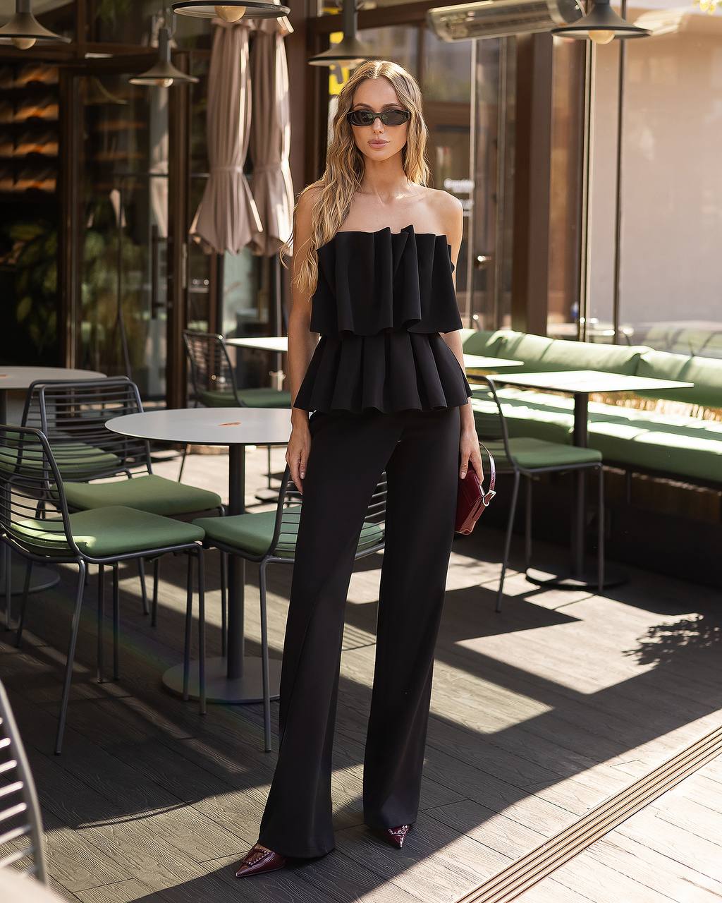 A woman stands on an outdoor patio wearing a black off-the-shoulder ruffled peplum top paired with black wide-leg trousers, dark sunglasses, and pointed shoes, with green-cushioned tables and chairs in the background.