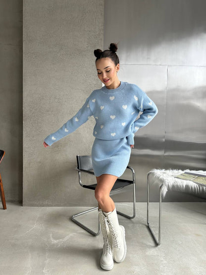 Woman wearing a light blue sweater and skirt set with heart patterns in a minimalistic room.