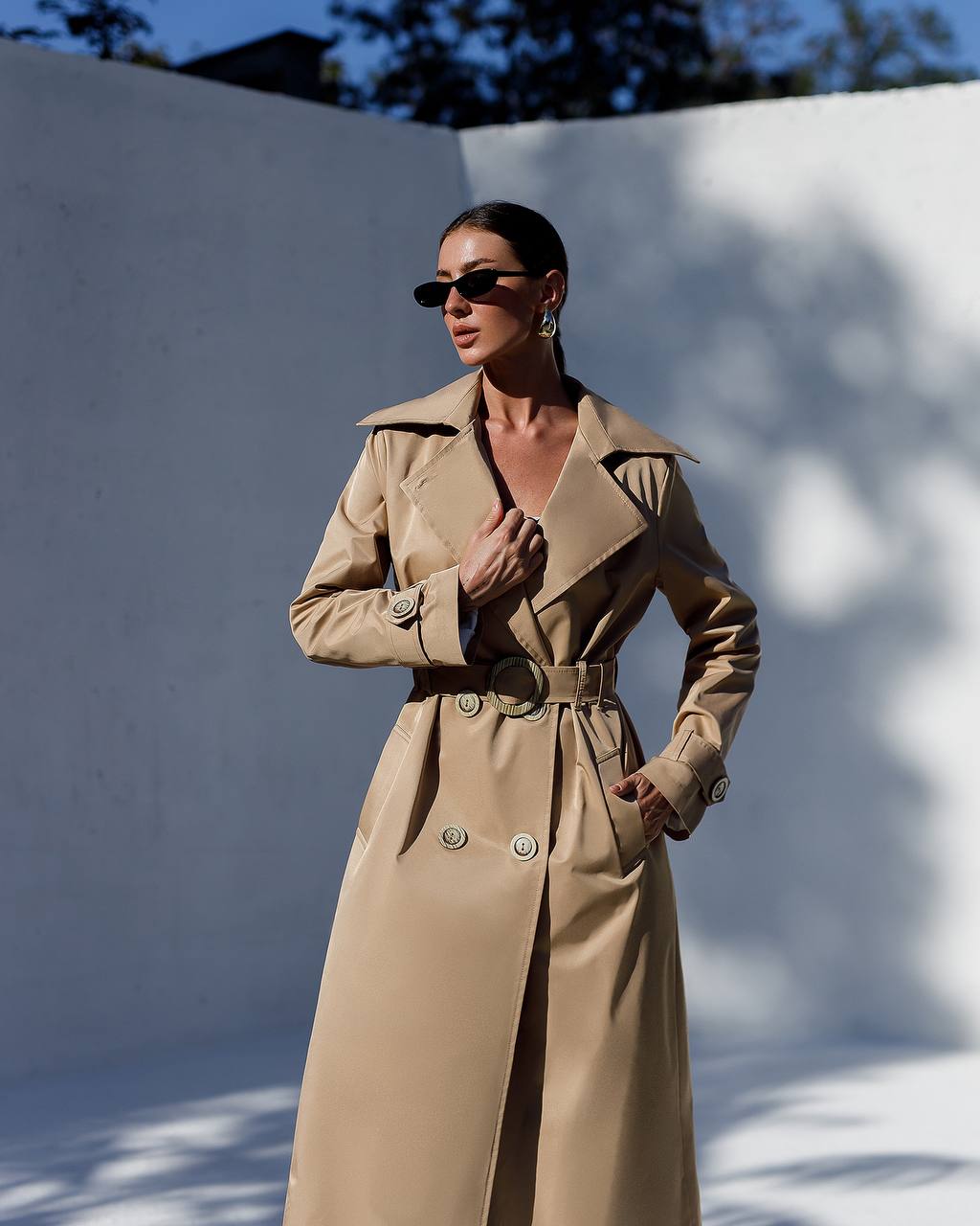 Woman wearing a beige trench coat standing against a white wall.