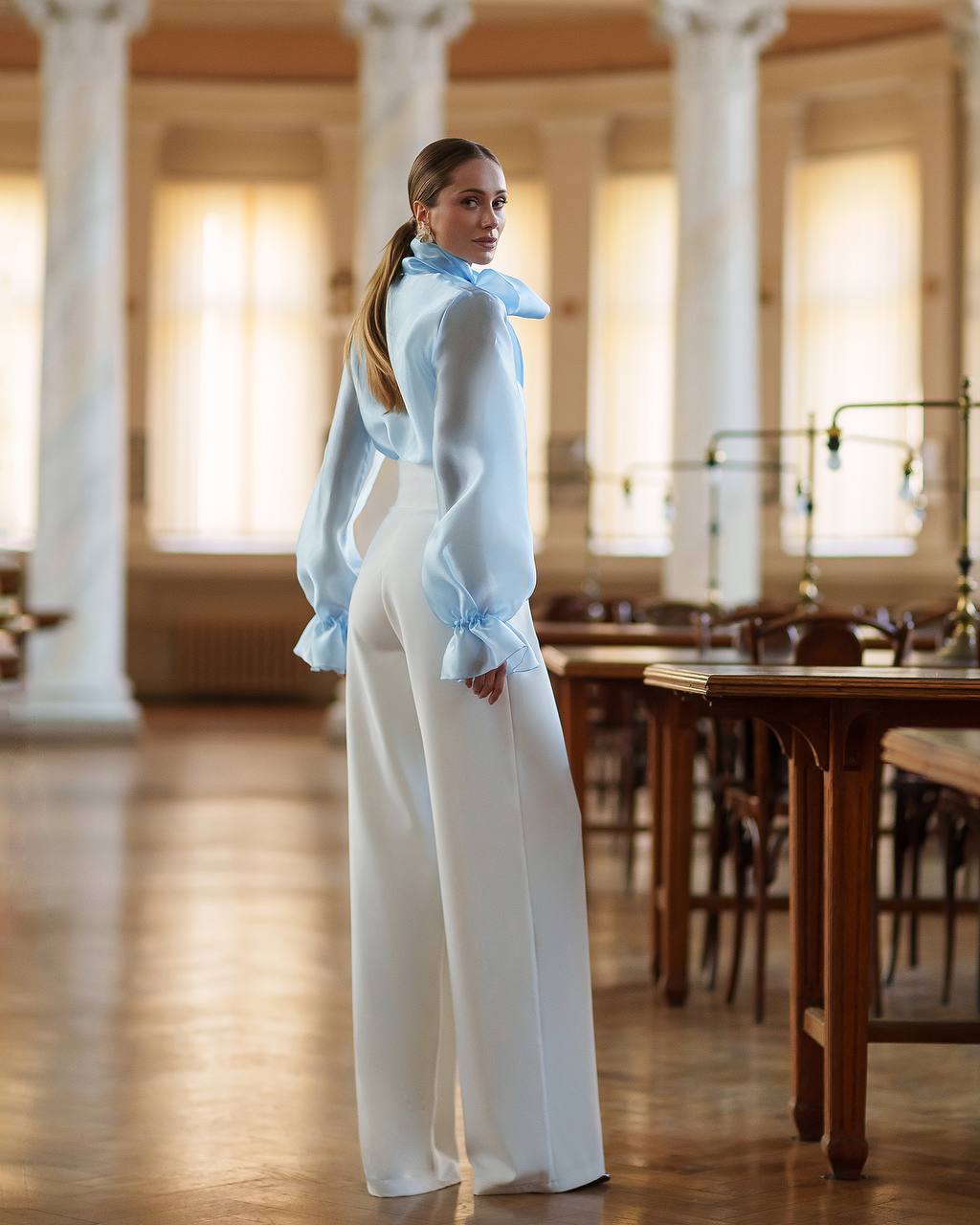 Woman in a light blue blouse and white pants standing in a large room with columns and wooden tables.