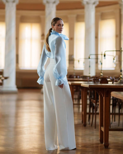 Woman in a light blue blouse and white pants standing in a large room with columns and wooden tables.