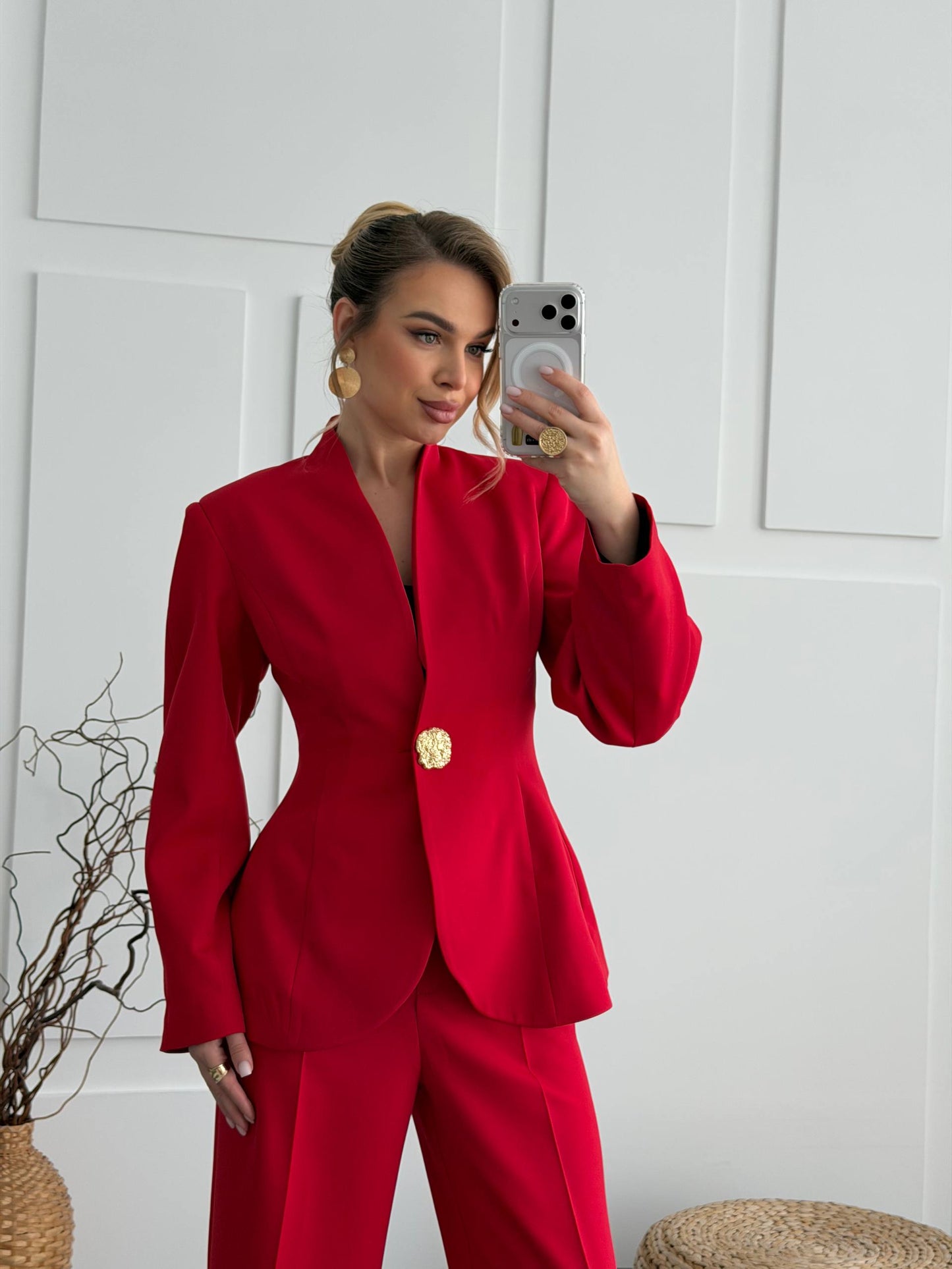 Woman taking a mirror selfie wearing a red suit with a gold button against a white wall.