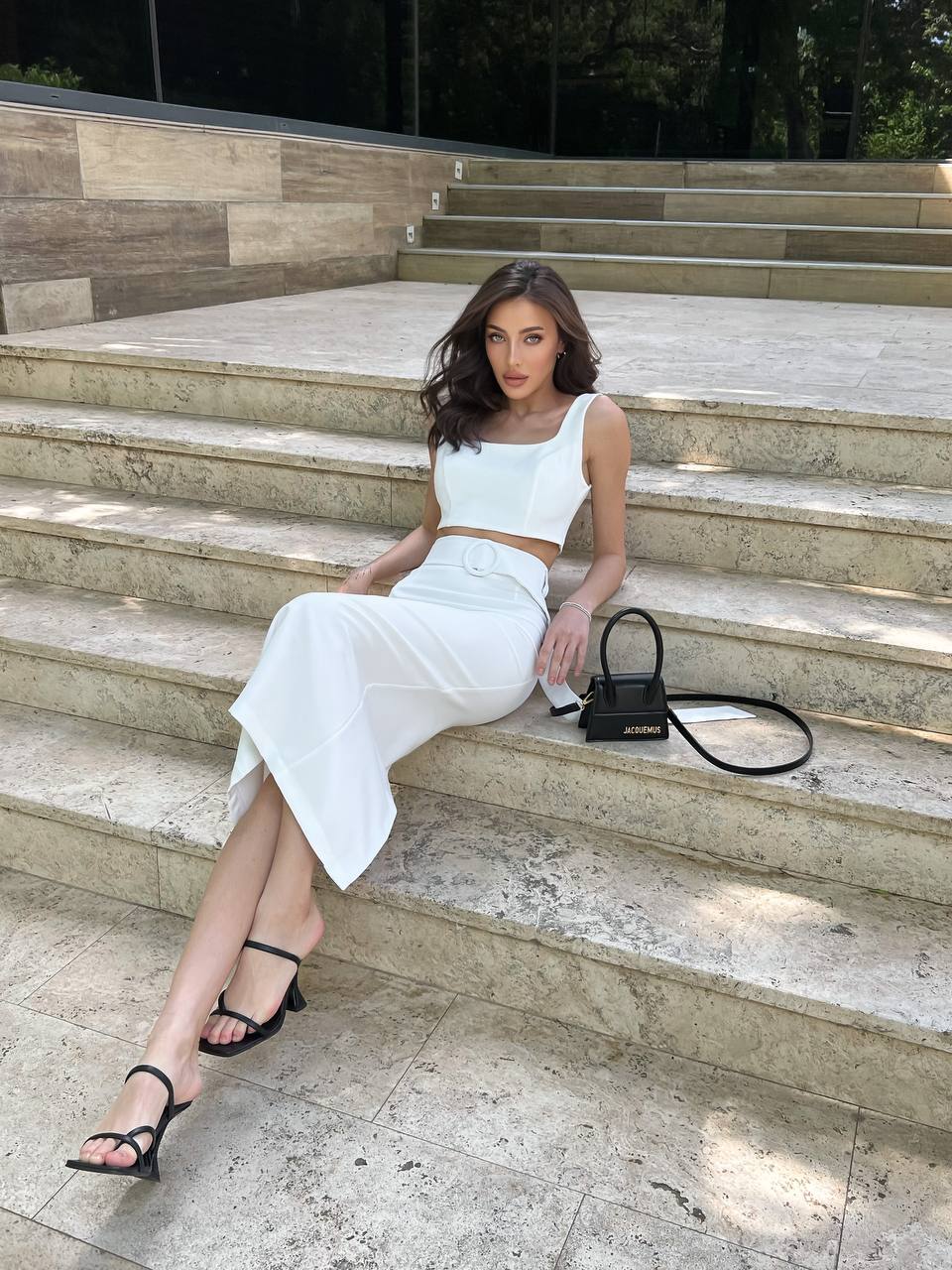 Woman in a white outfit sitting on stone steps with a black handbag.