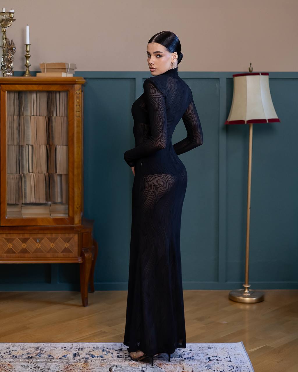 Woman in a long black dress standing in a room with a lamp and wooden cabinet.