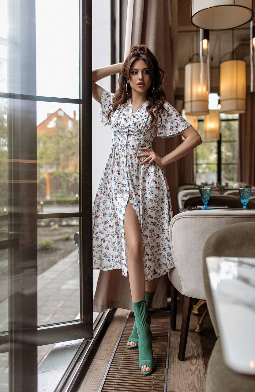 Woman in a floral dress standing in a modern interior setting with large windows.