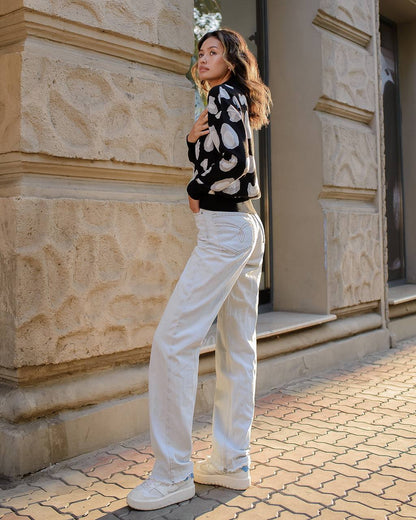 Woman in a black and white patterned sweater and white pants standing against a stone building.