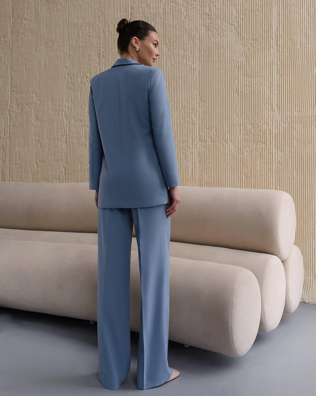 Woman in a blue suit standing in front of a beige sofa against a textured wall.