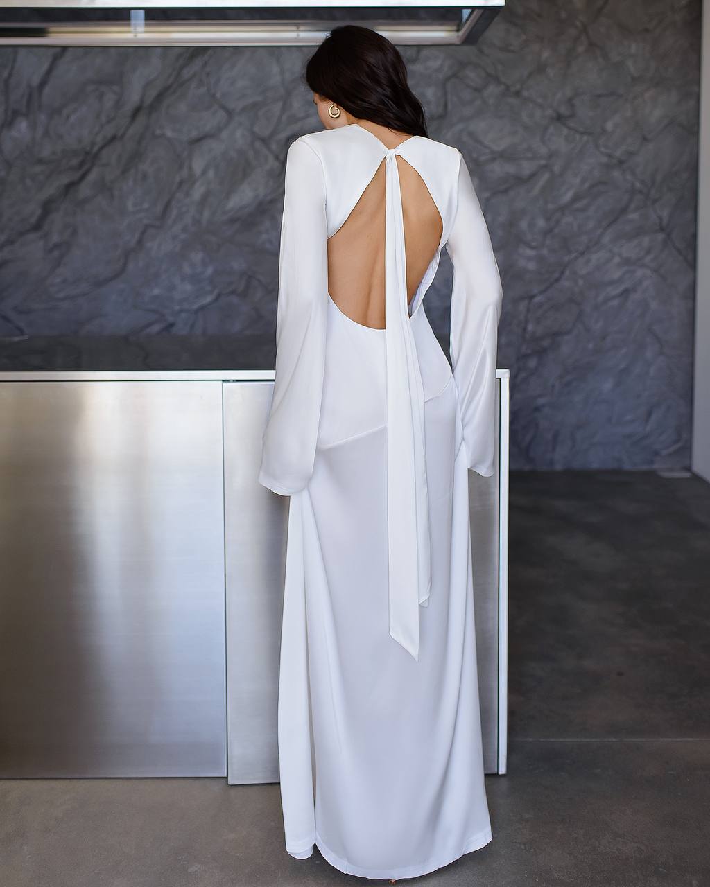 Woman wearing a white open-back dress standing against a dark textured wall.